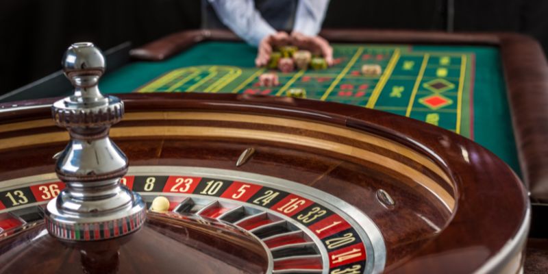 Untitled design (11) Roulette wheel and table with dealer pushing chips in background